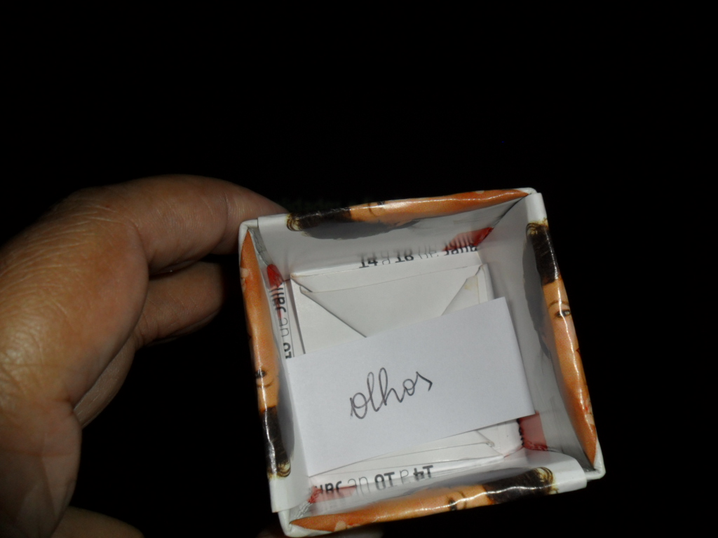 foto em fundo preto, uma mão segura uma caixa de papel, dentro dela 1 papel com a palavra olhos